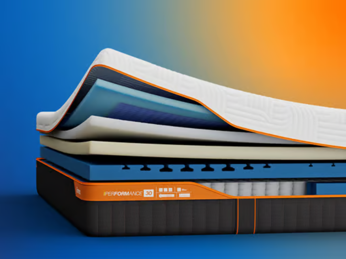 Emma Performance mattress corner showing a white quilted top, orange trim, and dark charcoal side panels with orange stitching, set against a blue-to-orange gradient background.