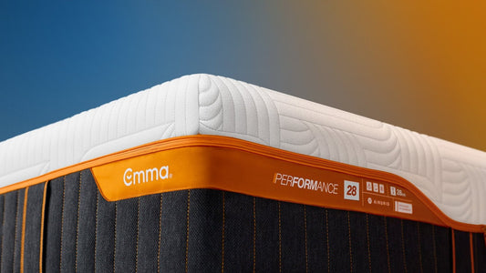 Emma Performance mattress corner showing a white quilted top, orange trim, and dark charcoal side panels with orange stitching, set against a blue-to-orange gradient background.