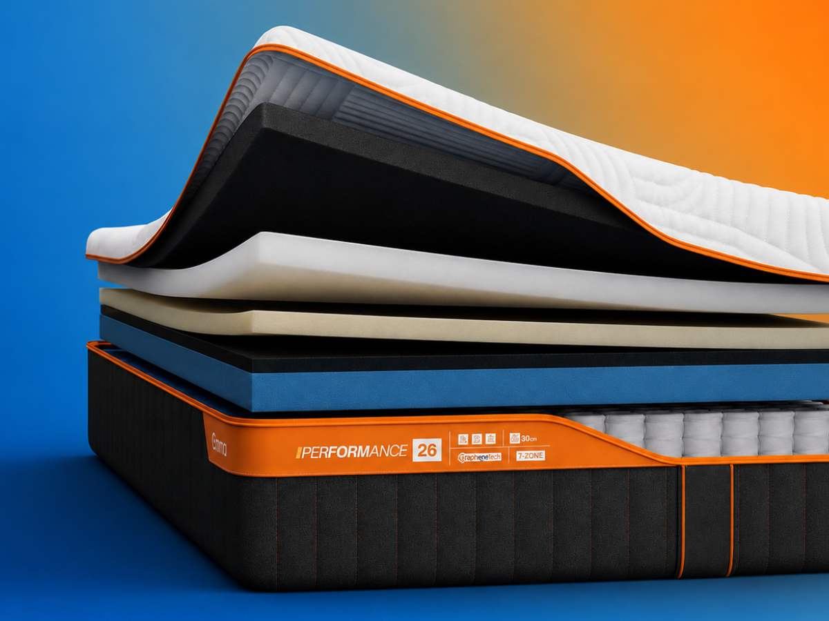 Emma Performance mattress corner showing a white quilted top, orange trim, and dark charcoal side panels with orange stitching, set against a blue-to-orange gradient background.